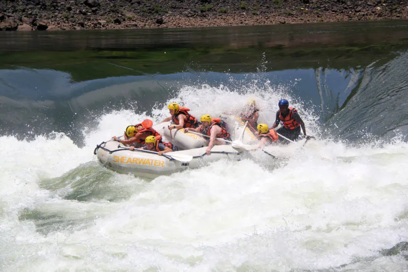 Group in yellow helmets paddling inflatable raft through rapids at Victoria Falls, splashing waves and rocky riverbanks.