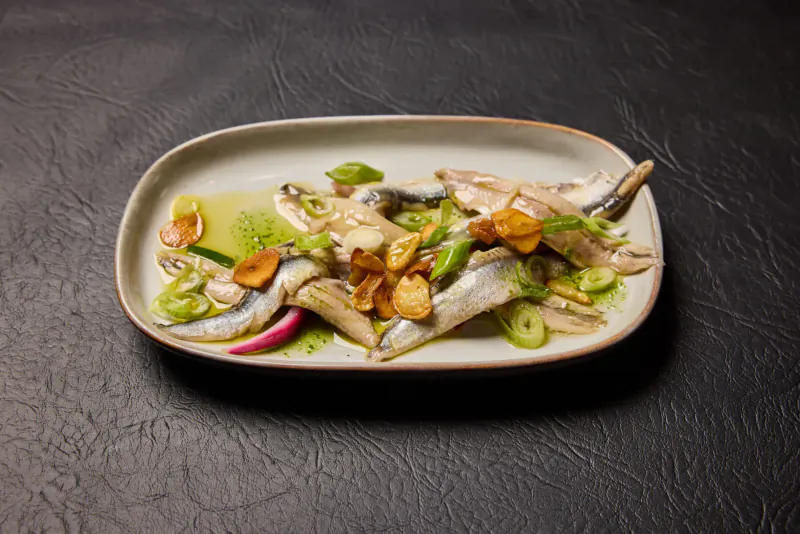 Oval plate of marinated sardines with garlic, green herbs, and oil on black surface