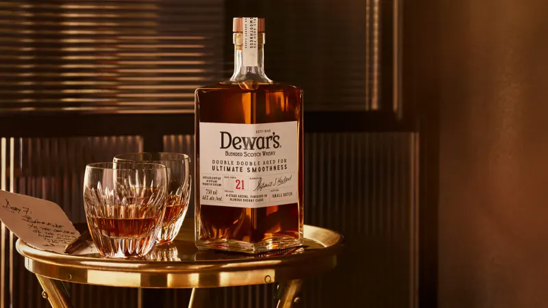 Dewar's 21 Year Old whisky bottle and glass of amber scotch on gold tray in warm setting