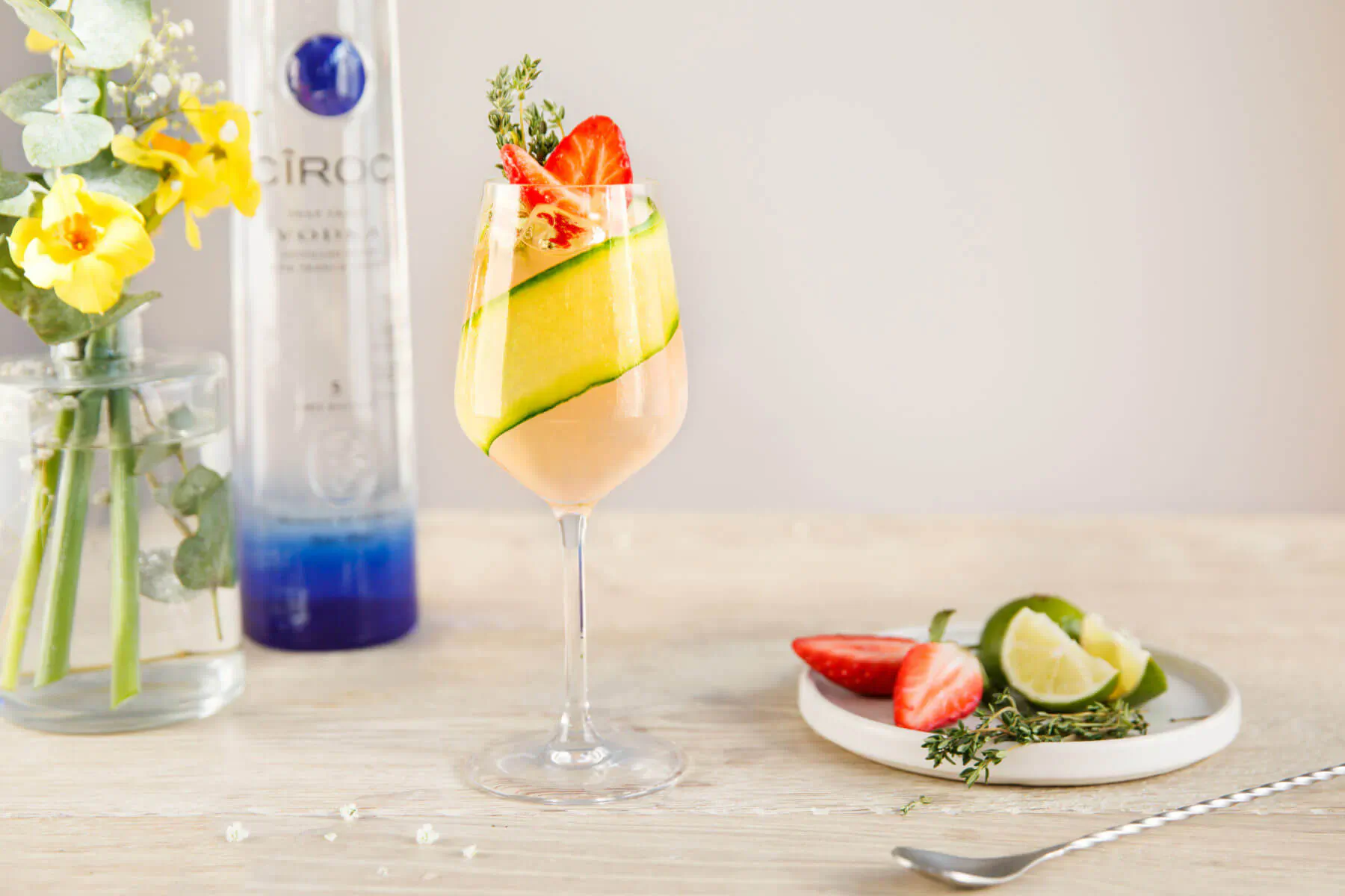 Cîroc in Bloom Spring cocktail in tall glass with strawberry, zucchini, thyme garnish, beside Cîroc bottle, yellow flowers, lime wedges