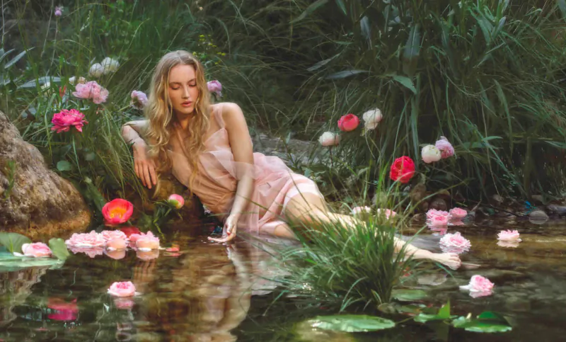 Blonde woman in pink dress reclining by lush green pond with pink flowers, water lilies, and rocks