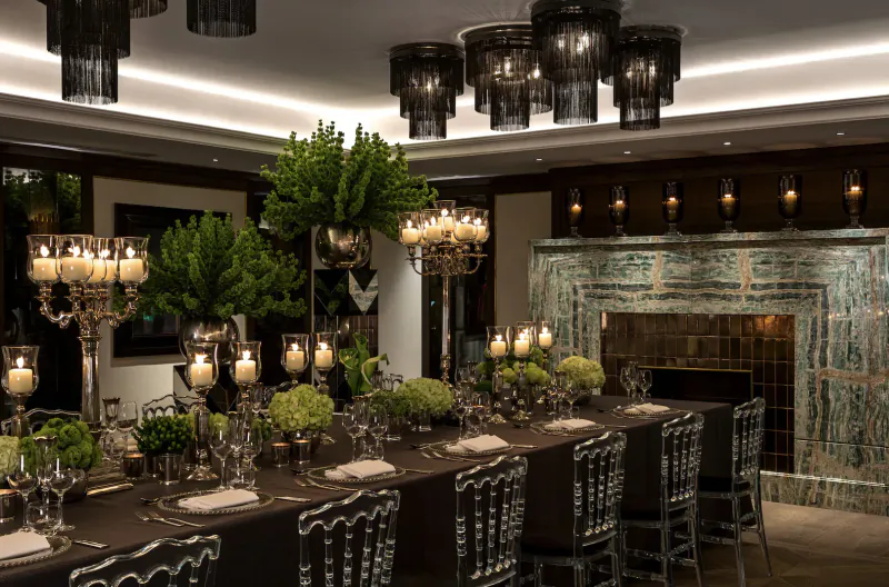 Elegant dining room at The May Fair Hotel with long table set for dinner, green plants, candelabras, fireplace, and black pendant lights.
