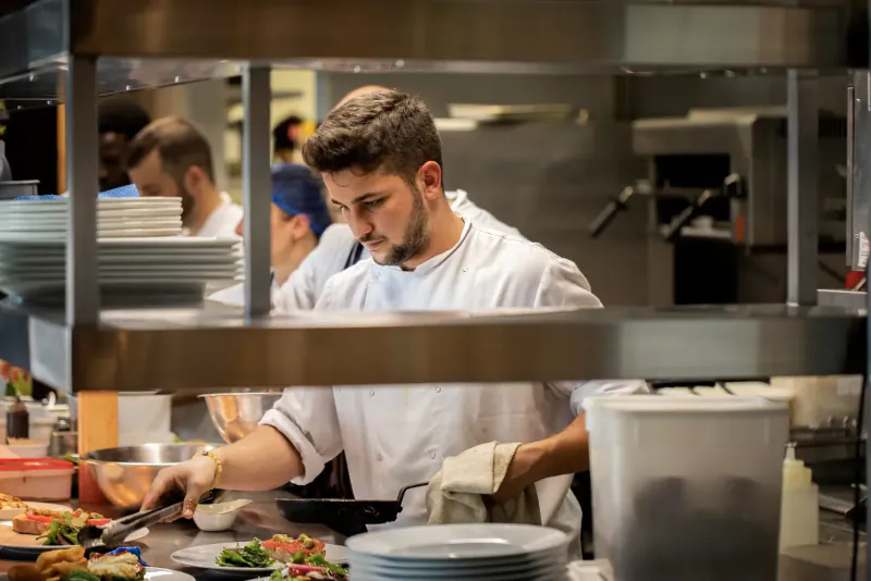 Bearded chef in white uniform scoops vibrant salad onto plate in busy commercial kitchen