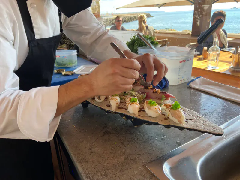 Chef in white uniform garnishing squid slices on wooden platter at beachside gastro counter with ocean view.