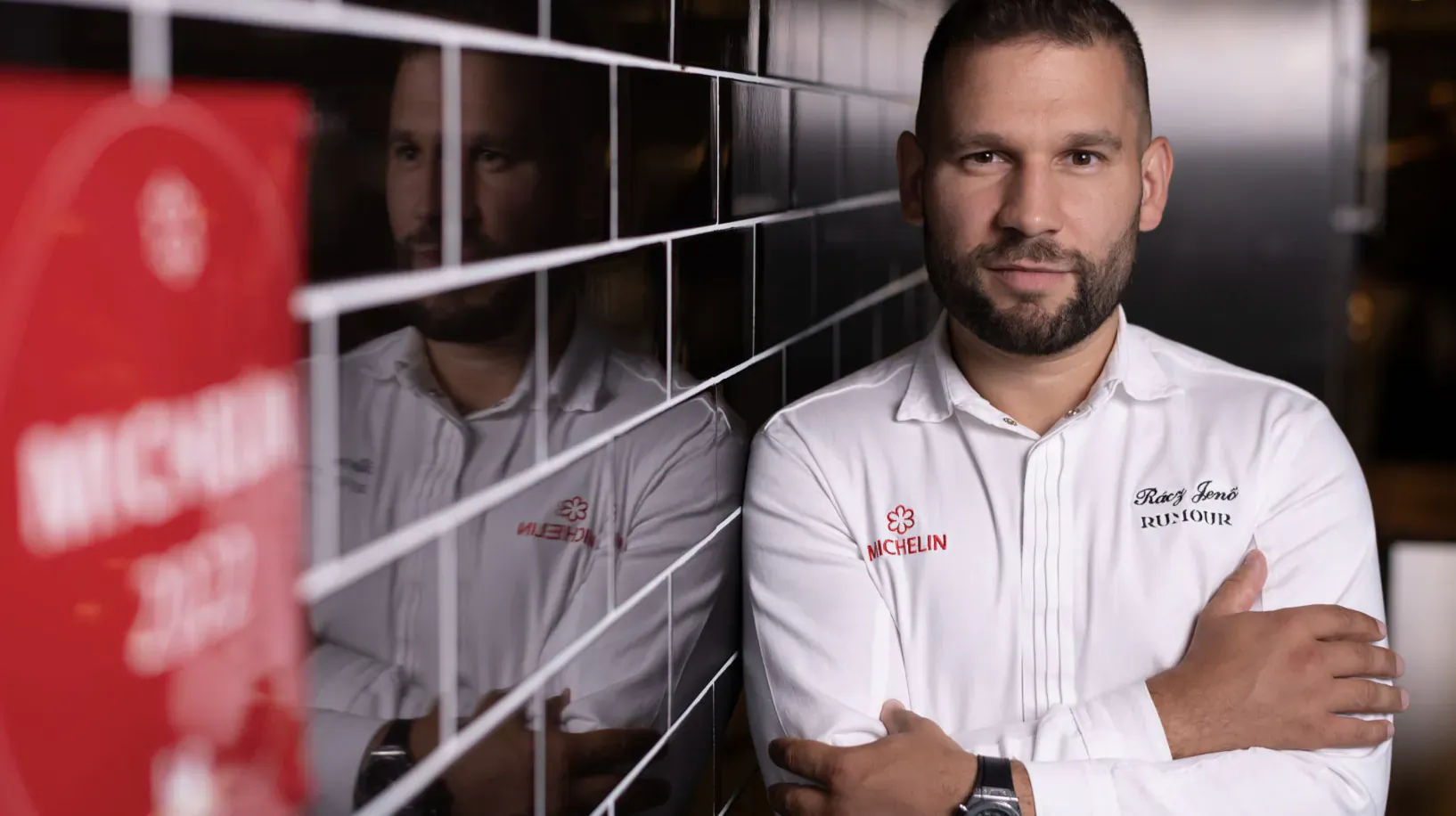 Michelin Star Chef Jeno Racz in white chef uniform with 'Black Salt' logo, arms crossed before tiled wall mirror reflection and red 'MICHELIN' sign.