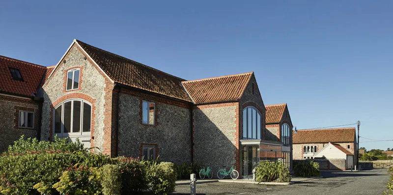 The Harper Norfolk: Stone barn-style country hotel with large arched windows, red-tiled roofs, bikes, hedges under blue sky.