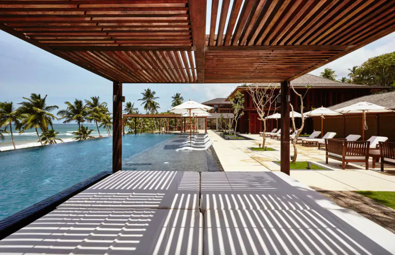 Infinity pool at Ani Private Resorts with wooden pergola, lounge chairs, umbrellas, palm trees, and beach view.