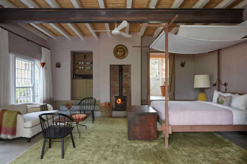 Cozy Franschhoek bedroom with pink four-poster bed, lit fireplace, wooden beams, green rug, and rattan chairs.