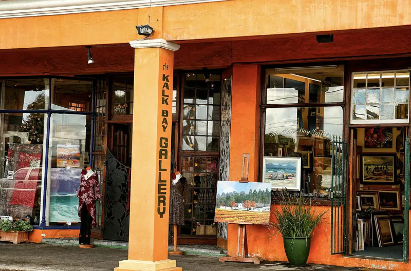 Orange facade of Walk-By Gallery in Kalk Bay, with large windows displaying paintings and sculptures outside.