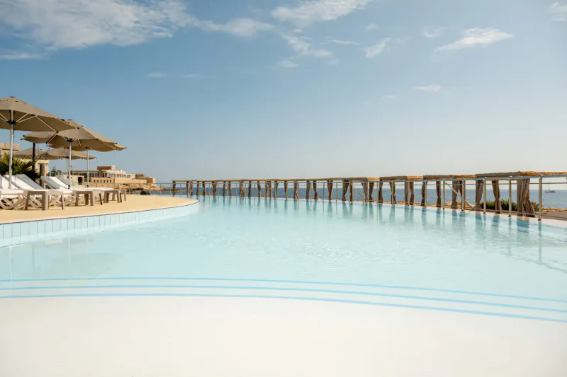 Infinity pool at Hilton Malta overlooking sea, with loungers, umbrellas, and wooden pier under blue sky.