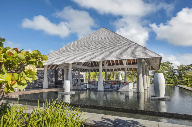 Open-air pavilion with peaked roof at Anantara Iko Mauritius Resort, surrounded by tropical plants and reflecting pool under blue sky.