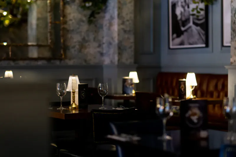 Elegant restaurant interior with candlelit tables, wine glasses, tufted banquettes, and framed artwork on ornate walls.