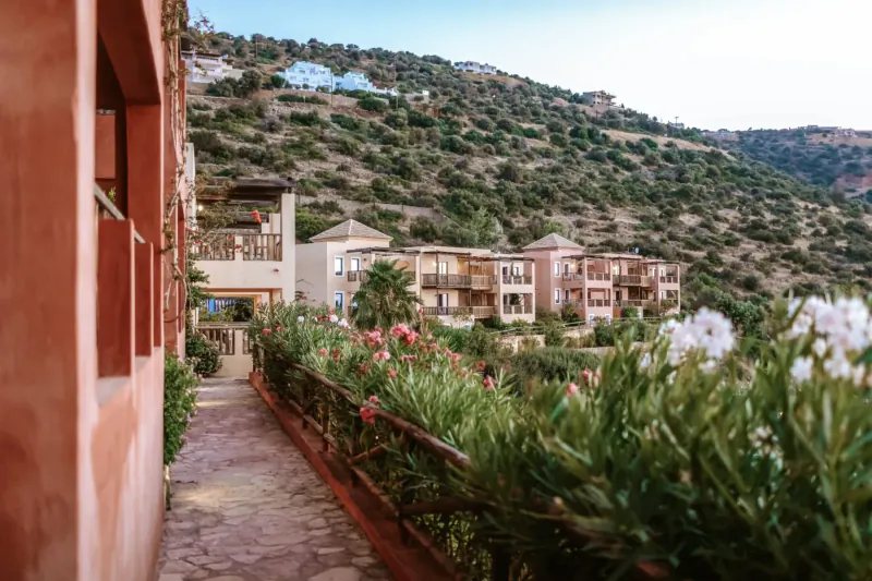 Stone pathway with pink flowers and lush greenery at Candia Park Village, overlooking terraced hillside villas in Crete