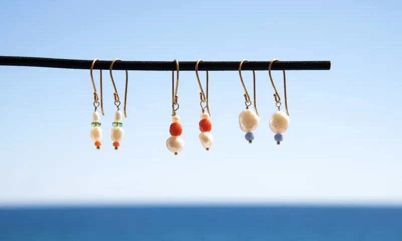 Assorted pearl drop earrings in white, orange, and blue beads hanging from a rod against blue sea backdrop