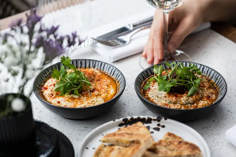Two bowls of hummus topped with greens and chili oil, pita bread, wine glass, on table with flowers at Mediterranean restaurant.