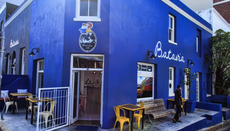 Blue 'Batavia' cafe exterior in Bo-Kaap with yellow chairs, tables, plants, and man walking outside.
