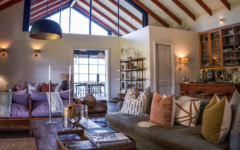 Spacious modern living room in Simbavati Cederberg Ridge retreat with wooden beams, plush gray sofa, colorful pillows, bar, and large windows.