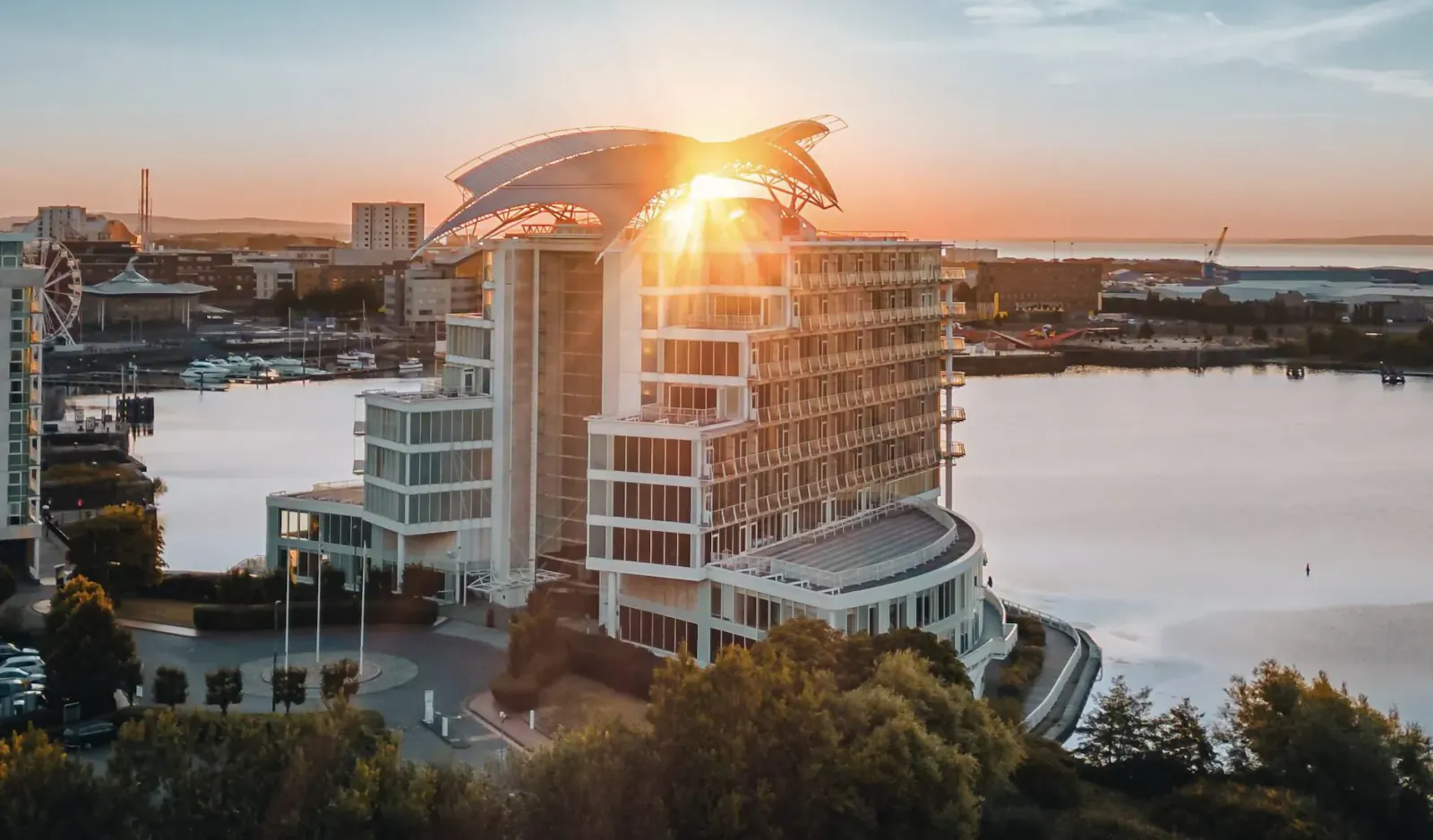 Aerial view of Voco St. David’s hotel with sail-like roof glowing in sunset over Cardiff's Tiger Bay waterfront and lake.