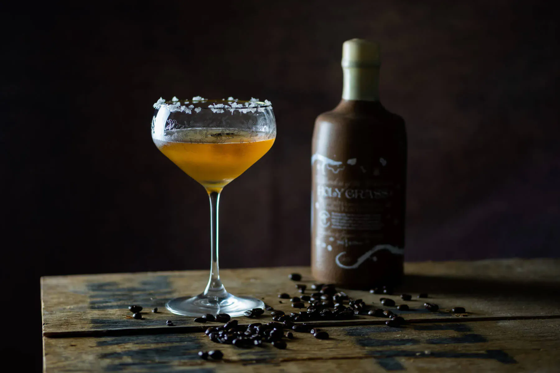Amber coffee vodka cocktail in salt-rimmed glass beside bottle labeled 'Kaffir Vodka' on coffee beans, dark setting.