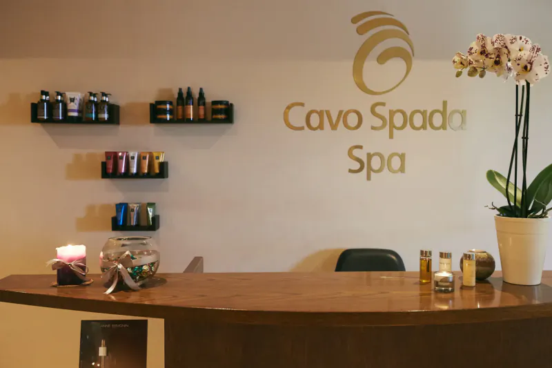 Cavo Spada Spa reception desk with beauty products on shelves, orchids, and gold logo on wall.