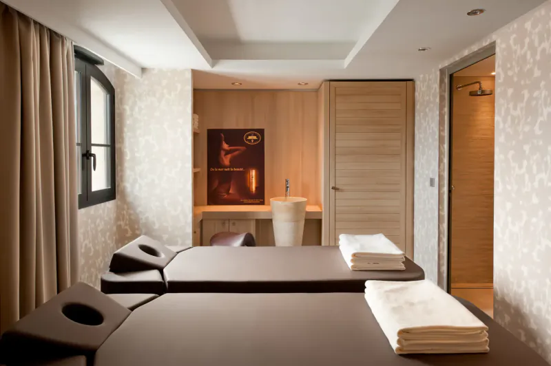Elegant spa room with two massage tables with towels, sink, shower, and wood accents.