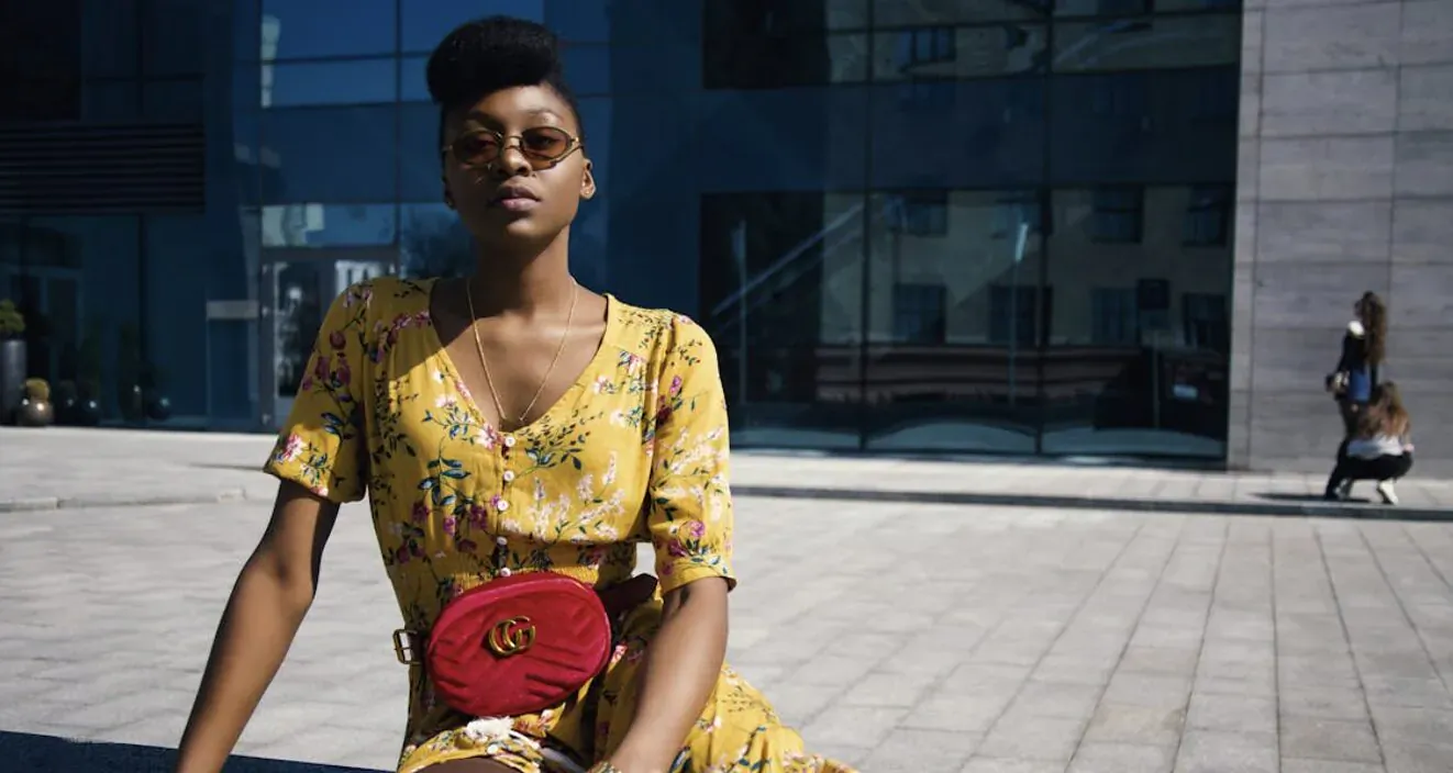 Black woman in yellow floral dress and red Gucci monogram bag sits on plaza bench before modern glass building