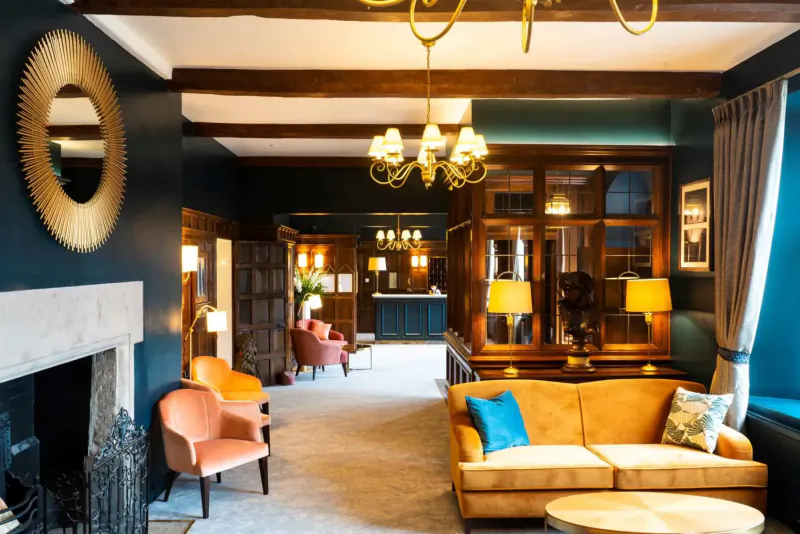 Elegant lounge at Billesley Manor Hotel & Spa with wooden beams, chandelier, orange armchairs, yellow sofa, fireplace, and bar.