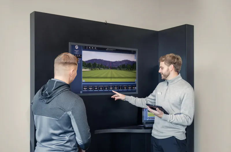 Two men in sportswear at a golf club fitting booth; one points to screen showing golf course landscape, other holds tablet.