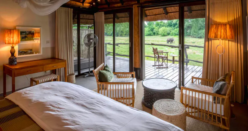 Spacious safari lodge bedroom with king bed, wooden chairs, fan, and veranda view of green bush and distant giraffe.