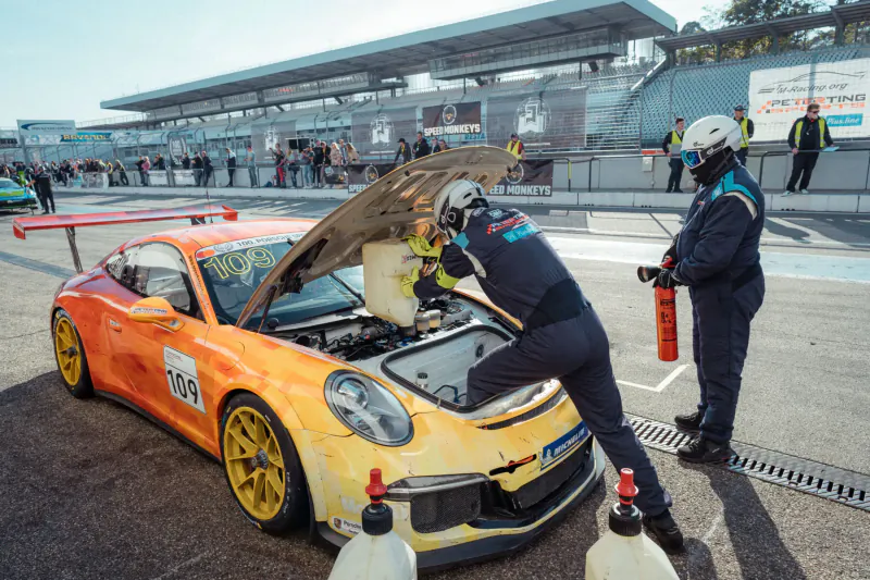 Two mechanics in blue suits and helmets work on orange #104 Porsche 911 GT3 race car with open hood at racetrack pit lane, fire extinguisher nearby.