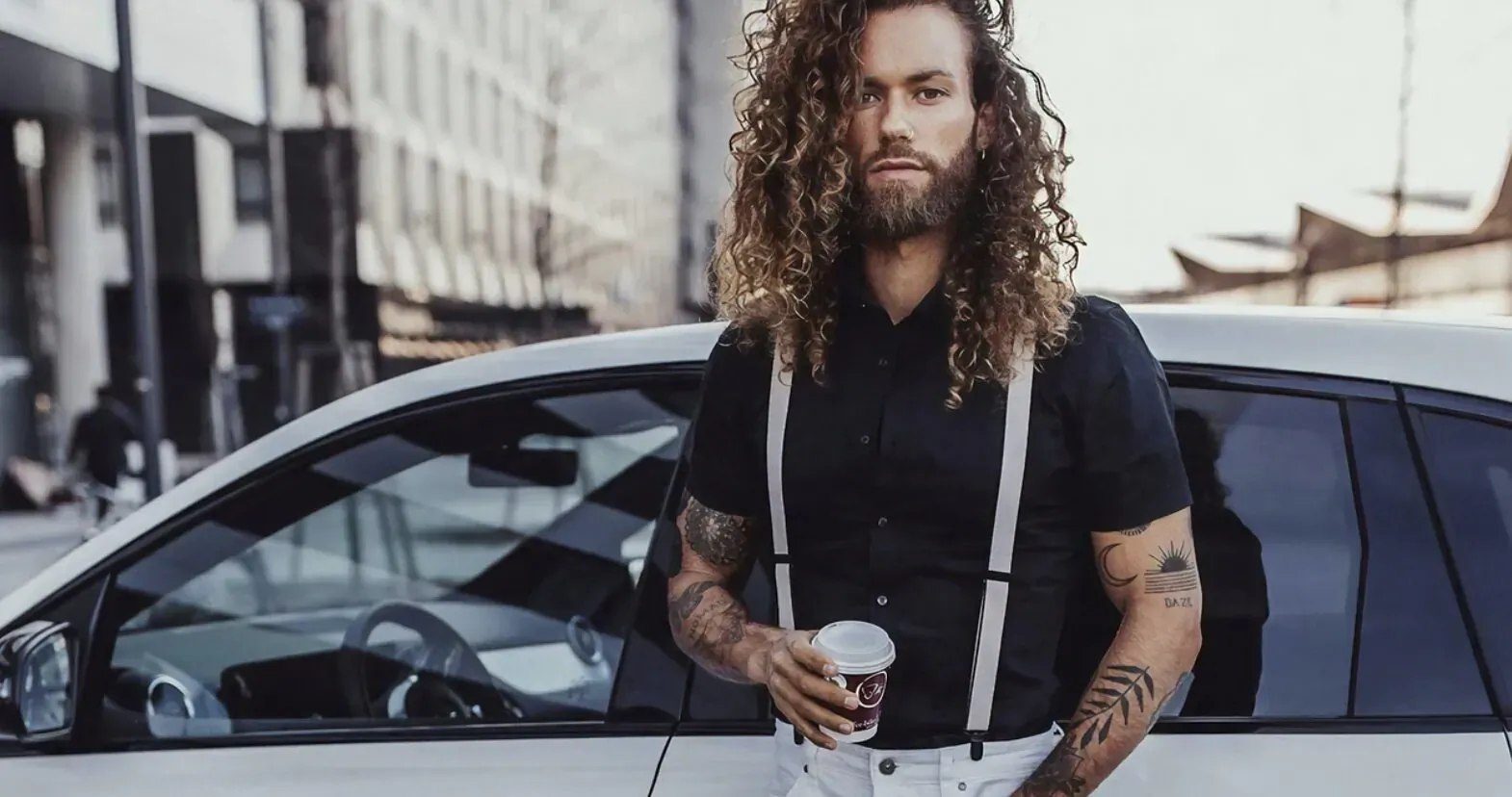 Long-haired bearded man with tattoos, in black shirt and suspenders, holding coffee cup beside white car in urban setting