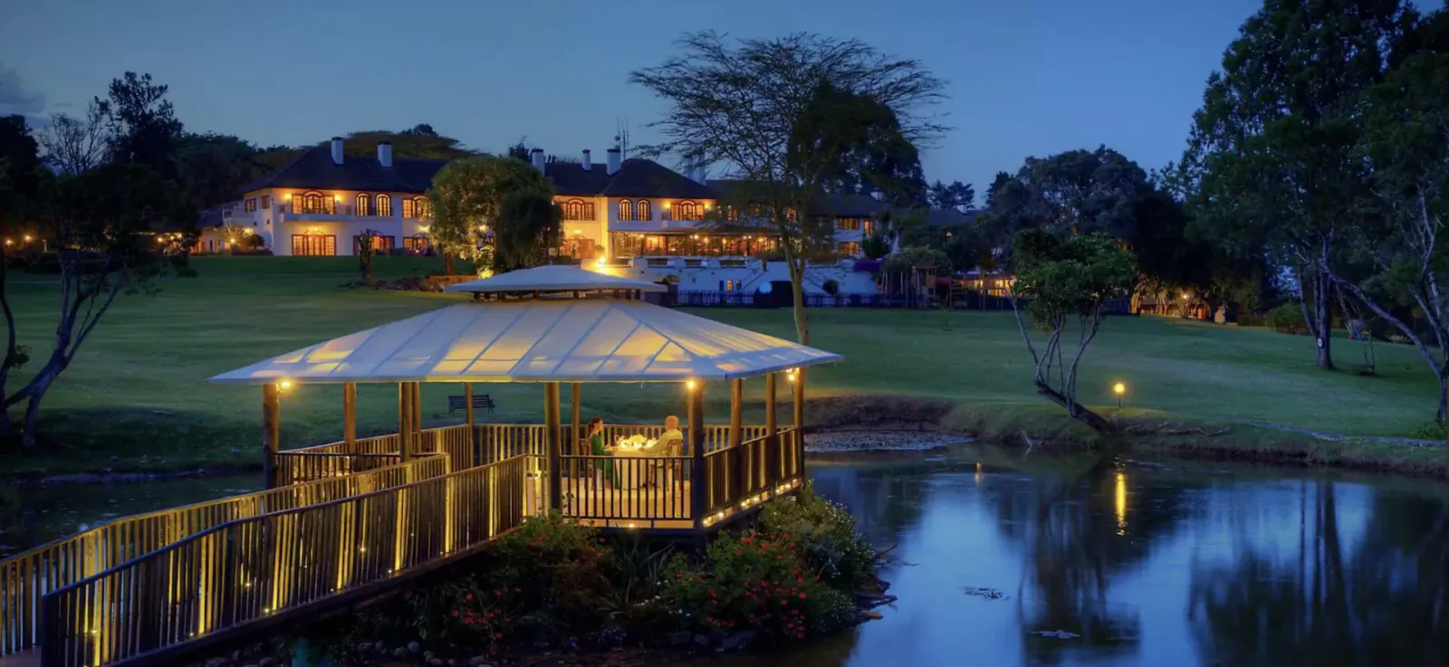 Twilight view of Fairmont Mount Kenya Safari Club: lit gazebo on bridge over pond, golf course, trees, and lodge in background