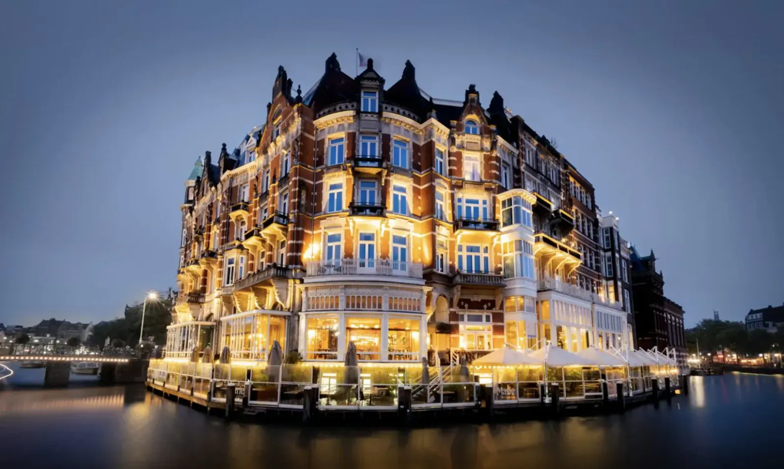 Lit-up ornate brick hotel on Amsterdam canal at night, corner view with reflections in water.