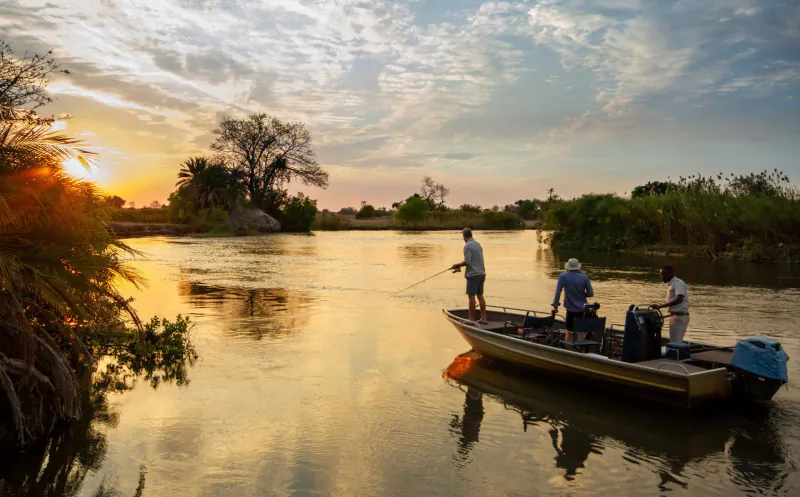 Three anglers fishing from a boat on the Okavango river at sunset, golden light and lush riverbanks.
