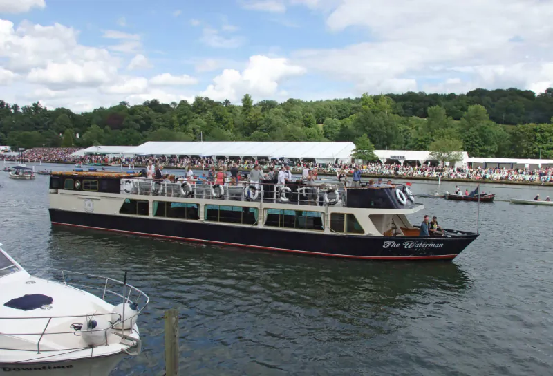 Hobbs of Henley passenger boat cruising on river amid regatta crowds, rowboats, and wooded shores under blue sky.