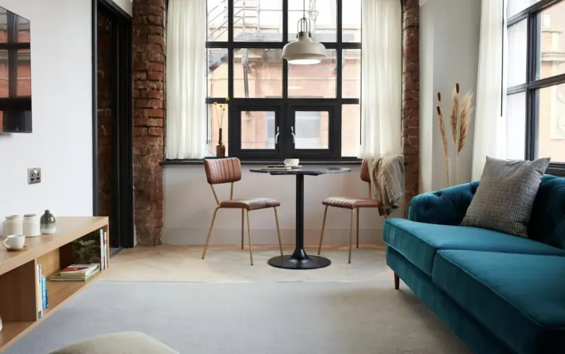 Modern living room in Leven Manchester with teal velvet sofa, black round table, leather chairs, brick walls, large windows.