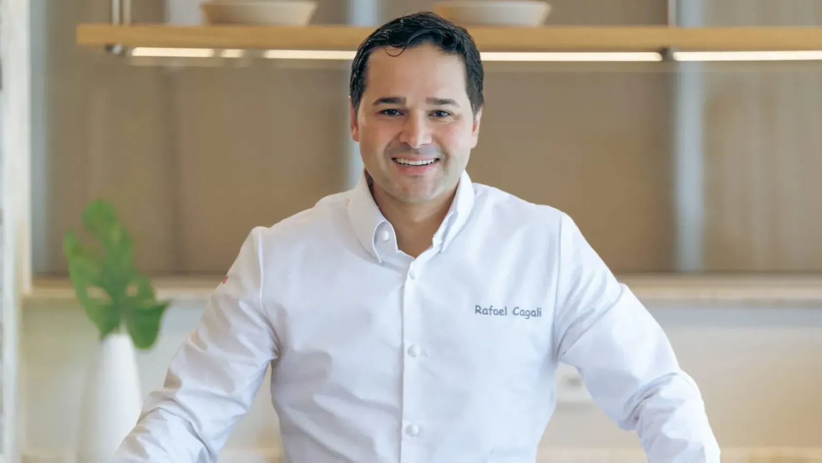 Smiling man in white chef shirt labeled 'Rafael Costa' stands in modern kitchen with plants and shelves.