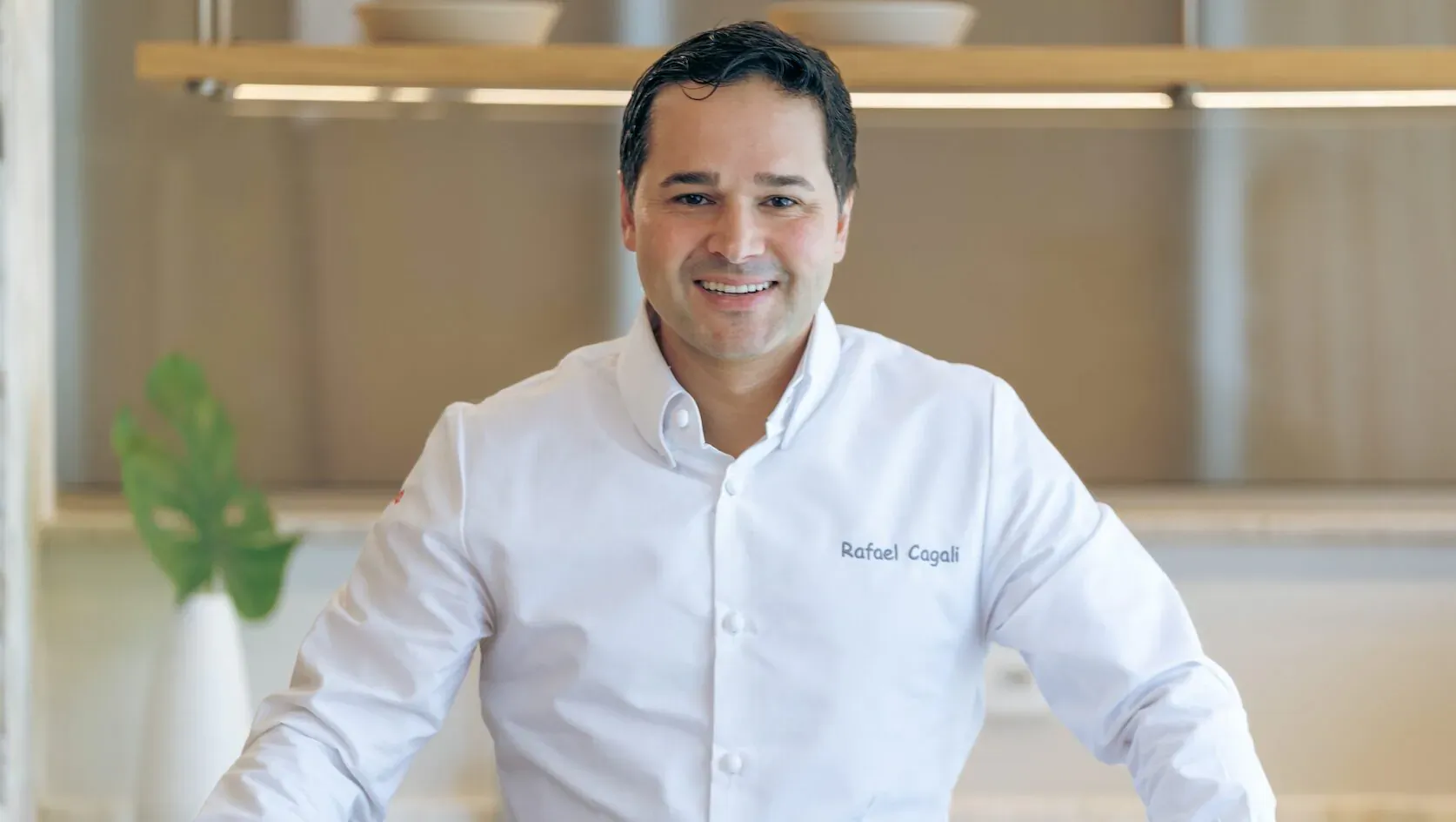 Smiling man in white chef shirt labeled 'Rafael Costa' stands in modern kitchen with plants and shelves.
