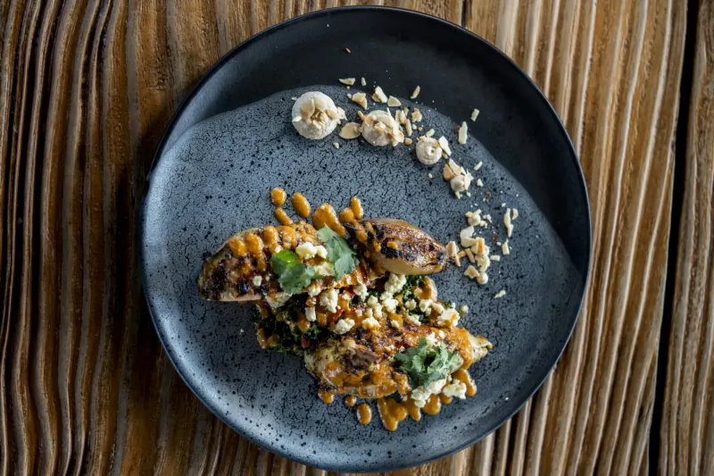 Close-up of grilled prawns in spicy sauce with chickpeas, herbs, and garnishes on black plate over wood.