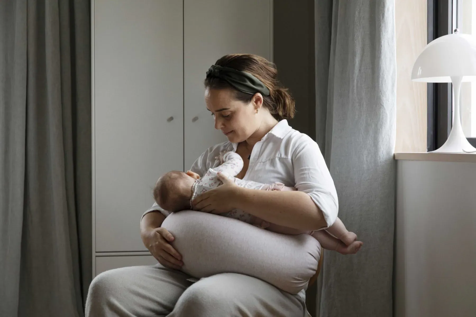 Mother breastfeeding newborn baby using nursing pillow in sunlit bedroom