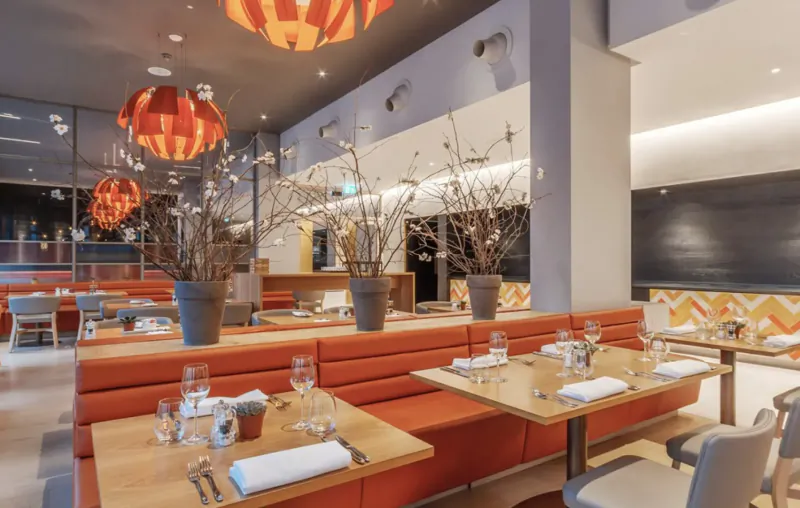 Modern Apex City of Bath Hotel restaurant interior with orange lamps, zigzag decor, large plants, and set tables.