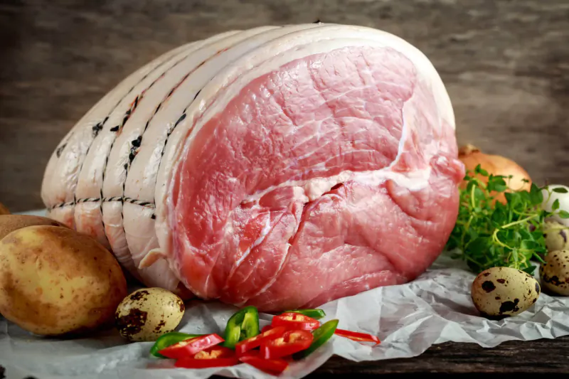 Large raw ham joint with scored rind on wooden surface, garnished with potatoes, quail eggs, herbs, and red chillies