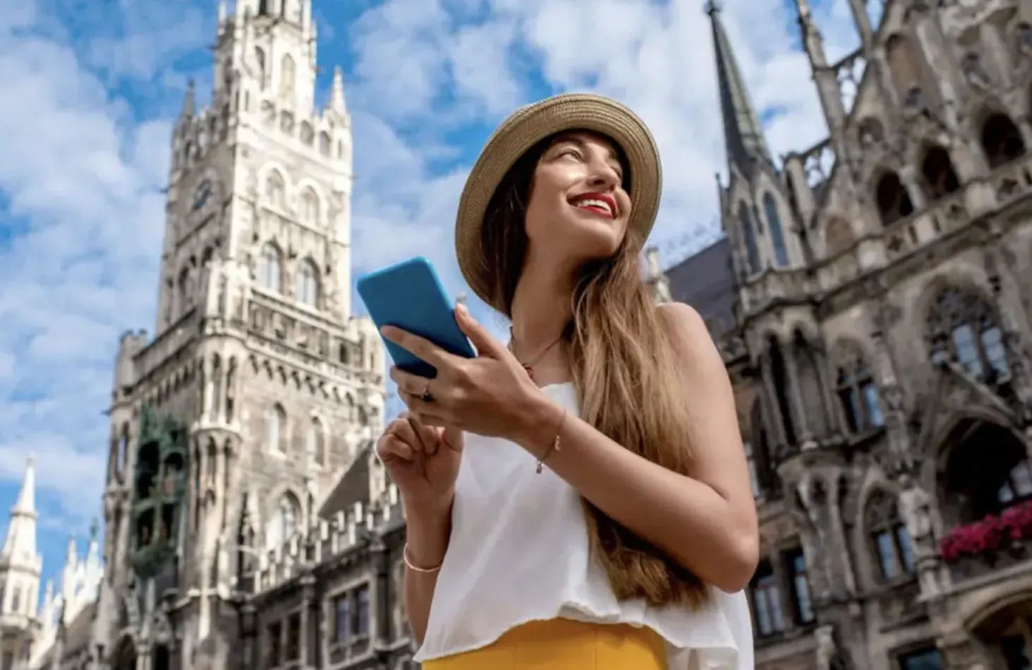 Smiling woman in beige hat and yellow skirt holds blue phone, smiling at Gothic cathedral in Europe under blue sky
