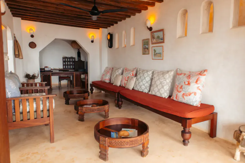 Serene Swahili lounge at Kijani Hotel Lamu with wooden furniture, orange pillows, arched walls, and ceiling fan.