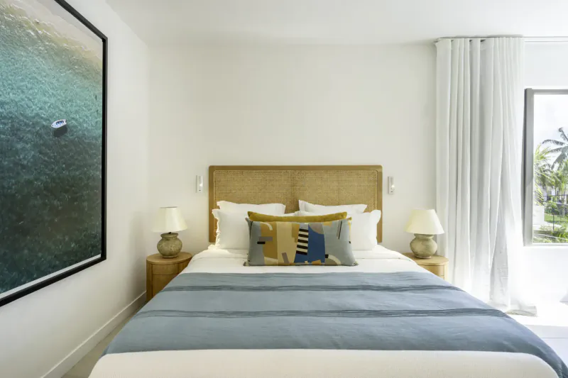 Spacious white bedroom in Villa Diamond, Mauritius, with tan rattan headboard, teal bedspread, geometric pillows, lamps, and large beach photo.