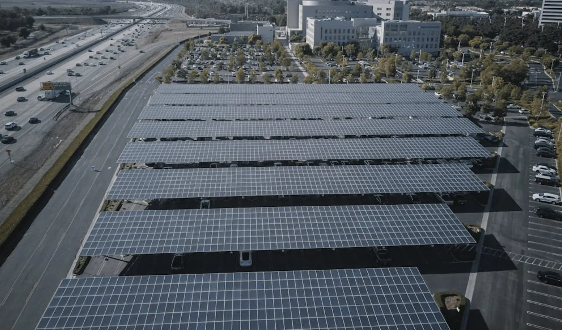 Aerial view of extensive solar panel carport array beside highway with traffic and buildings.