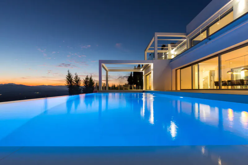 Modern white luxury house with infinity pool at twilight, overlooking mountains and sunset.