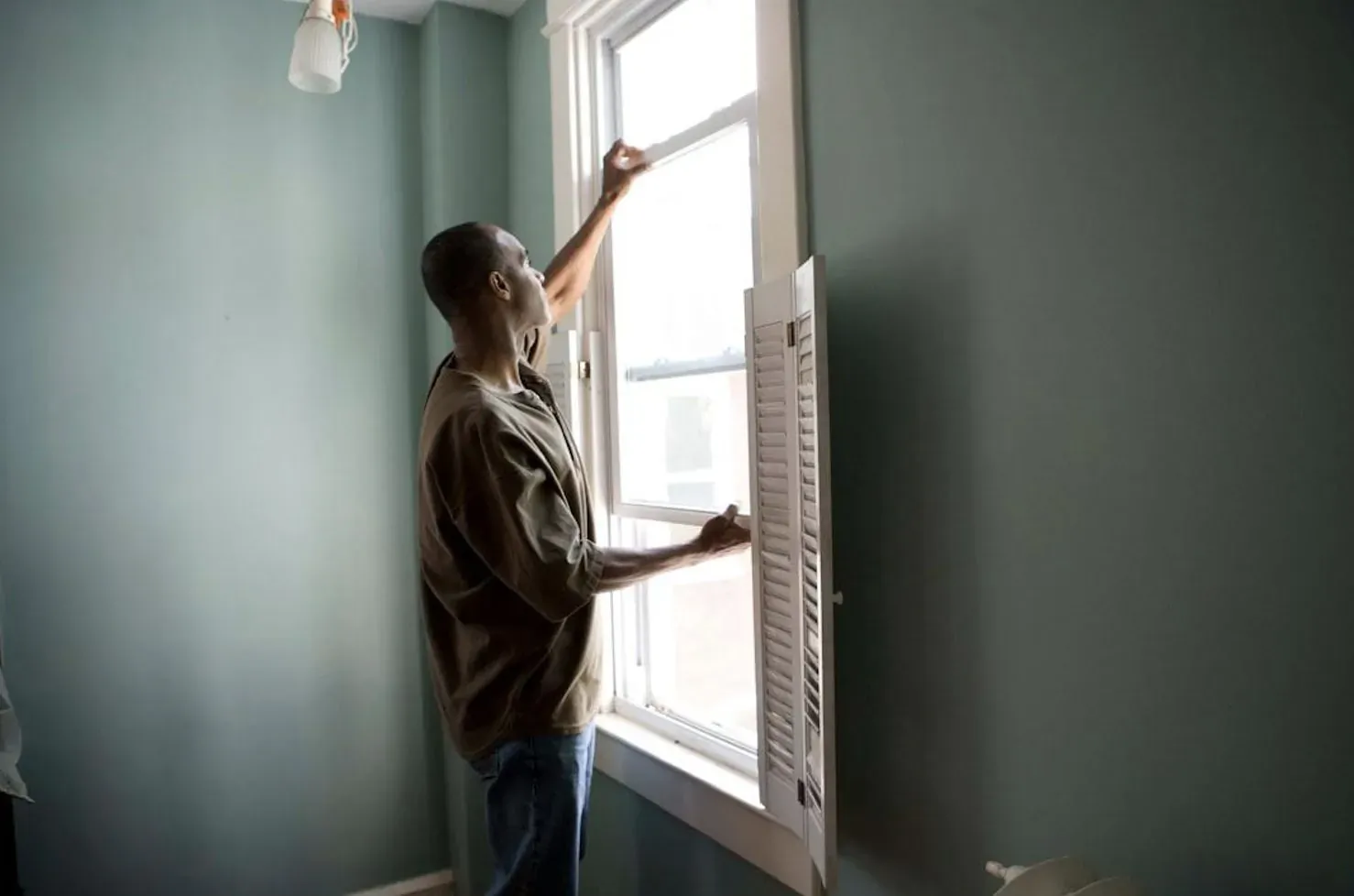 Man reaching up to adjust shutters on window in teal room while spraying for pests