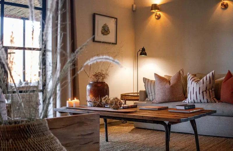 Cozy retreat living room with beige sofa, wooden coffee table, potted plants, candles, and warm lamp light by large window.