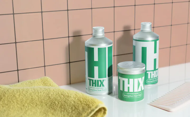 THIX caffeine hair products: green shampoo, silver conditioner bottles, tin jar on tiled shelf with towel and comb